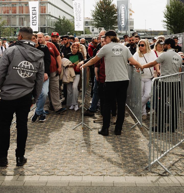 Einlass mit Sicherheitspersonal und Absperrgittern, dahinter wartende Besucher
